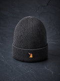 A ribbed beanie in charcoal standing on dark slate, the embroidered bird on the turn-up.
