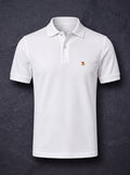 The St John Parker polo in white piqué on dark slate, the embroidered bird at the left chest.
